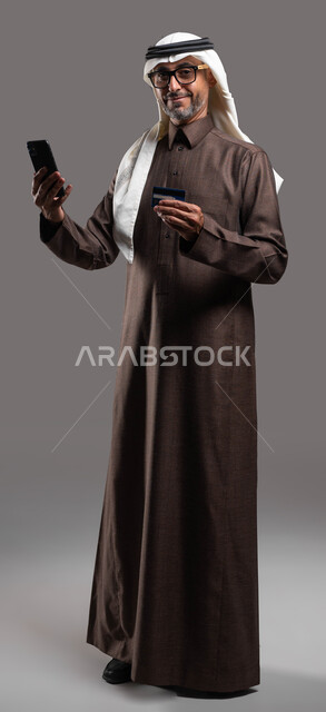 إمكانية الشراء وتسديد الفواتير من خلال برامج وتطبيقات الجوال، بورتريه لرجل شايب عربي خليجي سعودي يرتدي الثوب والغترة التقليدية يمسك بيده الهاتف المحمول وبطاقة الإئتمان البنكية ينظر إلى الكاميرا بإيماءات السرور بالتقنية الحديثة، صورة الجسم بالكامل، خلفية رمادية.