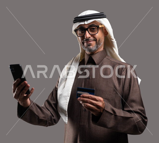 النظر إلى الكاميرا بإيماءات السرور باستخدام الوسائل الحديثة والمتطورة، الدفع الالكتروني عبر الانترنت، بورتريه مقرب لرجل شايب عربي خليجي سعودي مبتسم يرتدي الثوب والغترة التقليدية يمسك بيده الهاتف المحمول وبطاقة الإئتمان البنكية، خلفية رمادية.