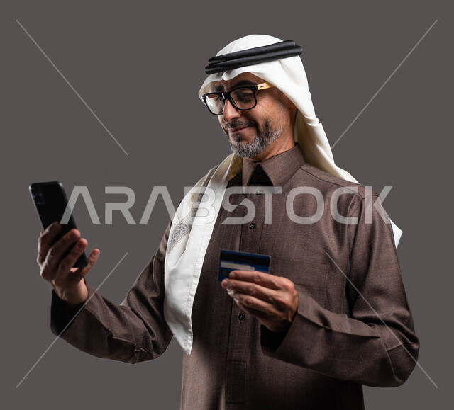الدفع الالكتروني عبر الانترنت، بورتريه مقرب لرجل شايب عربي خليجي سعودي يرتدي الثوب والغترة التقليدية يمسك بيده الهاتف المحمول وبطاقة الإئتمان البنكية بإيماءات السرور باستخدام وسائل التكنولوجيا، خلفية رمادية.
