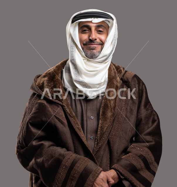 Feeling the bitter cold in the winter, looking at the camera with gestures of pleasure and self-confidence, the importance of wearing warm clothes for the elderly in winter conditions, a close-up portrait of a smiling Saudi Arabian Gulf gentleman wearing a traditional fur and ghutra, gray background.
