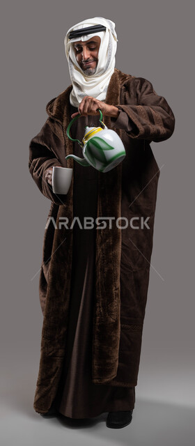 الاستمتاع بتناول المشروبات والأعشاب الساخنة في الشتاء، ارتداء الملابس الدافئة في الأجواء الشتوية، بورتريه لسيد عربي سعودي خليجي يرتدي الفروة والغترة التقليدية يقوم بسكب الشاي الساخن، صورة لكامل الجسم، خلفية رمادية.