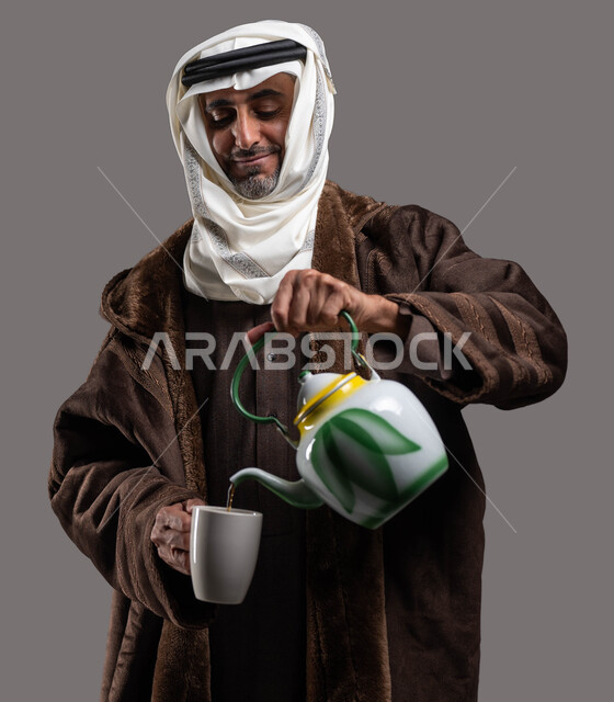 أهمية ارتداء الملابس الدافئة في أجواء فصل الشتاء، الشعور بالمتعة والدفء بتناول المشروبات والأعشاب الشتوية الساخنة، بورتريه مقرب لسيد عربي سعودي خليجي مبتسم يرتدي الفروة والغترة التقليدية يقوم بسكب الشاي الساخن، خلفية رمادية.