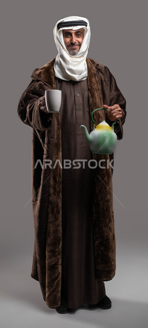 Looking at the camera with gestures of pleasure and welcome, drinking hot drinks and herbs in the winter, wearing warm clothes in the winter weather, a portrait of a Saudi Arabian Gulf gentleman wearing a traditional fur and ghutra, holding a teapot and a glass cup in his hand, a full-body portrait, gray background.