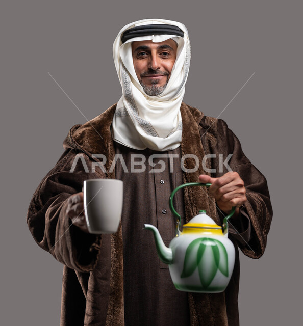 تناول كبار السن المشروبات الساخنة لتدفئة الجسم في الشتاء، النظر إلى الكاميرا بإيماءات السرور، ارتداء الملابس الدافئة في الأجواء الشتوية، بورتريه مقرب لسيد عربي سعودي خليجي مبتسم يرتدي الفروة والغترة التقليدية يمسك بيده ابريق الشاي والكوب الزجاجي، خلفية رمادية.
