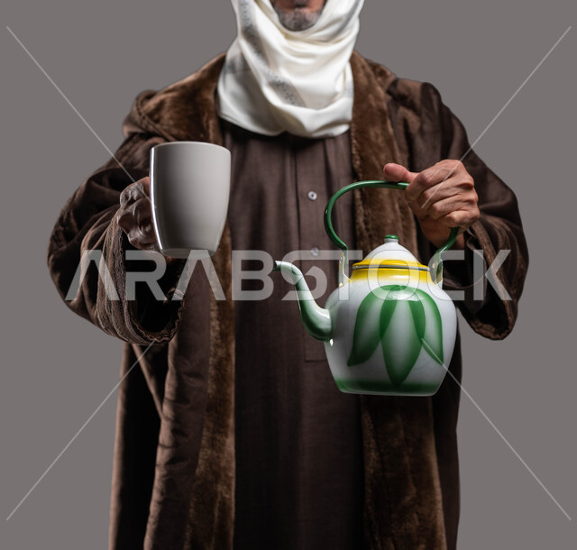 تناول المشروبات والأعشاب الساخنة في الشتاء، حسن الضيافة والاستقبال، ارتداء الملابس الدافئة في الأجواء الشتوية، بورتريه مقرب لسيد عربي سعودي خليجي يرتدي الفروة والغترة التقليدية يمسك بيده ابريق الشاي والكوب الزجاجي، خلفية رمادية.