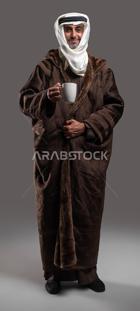 النظر إلى الكاميرا بإيماءات السرور، ارتداء الملابس الدافئة في الأجواء الشتوية، تدفئة جسم كبار السن في فصل الشتاء بتناول المشروبات الساخنة، بورتريه لسيد عربي سعودي خليجي مبتسم يرتدي الفروة والغترة التقليدية يمسك بيده كوب من الشاي الساخن، صورة الجسم بالكامل، خلفية رمادية.