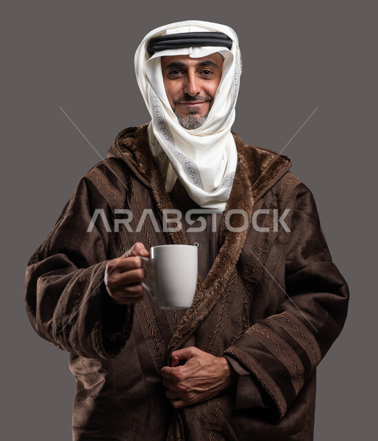 تناول كبار السن المشروبات الساخنة لتدفئة الجسم في الشتاء، النظر إلى الكاميرا بإيماءات السرور، ارتداء الملابس الدافئة في الأجواء الشتوية، بورتريه مقرب لسيد عربي سعودي خليجي مبتسم يرتدي الفروة والغترة التقليدية يمسك بيده كوب من الشاي الساخن، خلفية رمادية.