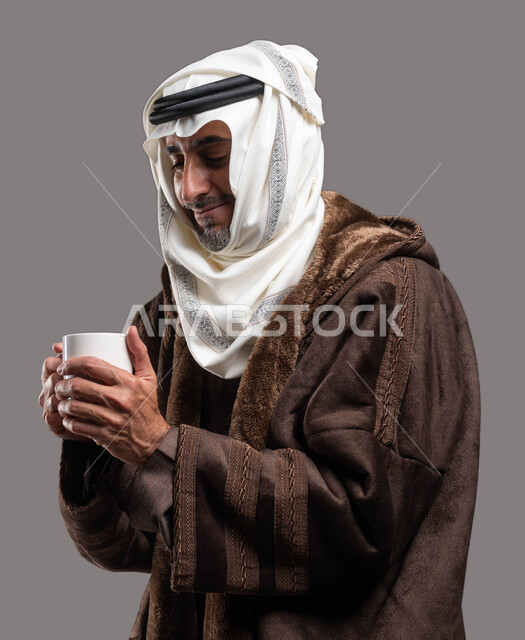 تدفئة جسم كبار السن في فصل الشتاء بتناول المشروبات الساخنة، ارتداء الملابس الدافئة في الأجواء الشتوية، بورتريه مقرب لسيد عربي سعودي خليجي يرتدي الفروة والغترة التقليدية يمسك بيده كوب من الشاي الساخن، خلفية رمادية.