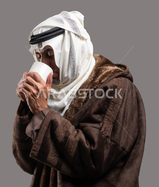 العناية بصحة كبار السن في فصل الشتاء بارتداء الملابس الدافئة، الاستمتاع بالمشروبات الساخنة في الأجواء الشتوية، بورتريه مقرب لسيد عربي سعودي خليجي يرتدي الفروة والغترة التقليدية يقوم بشرب كوب من الشاي الساخن، خلفية رمادية.
