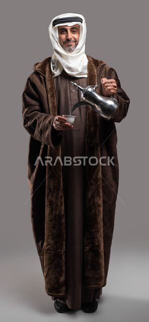 أساليب عريقة في كرم الضيافة، النظر إلى الكاميرا بإيماءات السرور والترحيب، حسن استقبال وإكرام الضيوف، بورتريه لسيد عربي سعودي خليجي مبتسم يرتدي الفروة والغترة التقليدية يقوم بسكب القهوة العربية الساخنة، صورة الجسم بالكامل، خلفية رمادية.