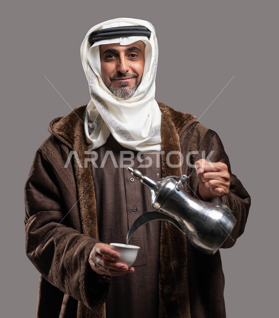 النظر إلى الكاميرا بإيماءات السرور والترحيب، استقبال وإكرام الضيوف وفق العادات والتقاليد الأصيلة المتوارثة، أساليب عريقة في كرم الضيافة، بورتريه مقرب لسيد عربي سعودي خليجي مبتسم يرتدي الفروة والغترة التقليدية يقوم بسكب القهوة العربية الساخنة، خلفية رمادية.