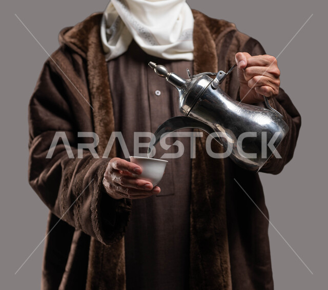 حسن استقبال وإكرام الضيوف وفق العادات والتقاليد الأصيلة المتوارثة، بورتريه مقرب لسيد عربي سعودي خليجي يرتدي الفروة والغترة التقليدية يقوم بسكب القهوة العربية ، الاستمتاع بتناول المشروبات الساخنة في الشتاء، خلفية رمادية.
