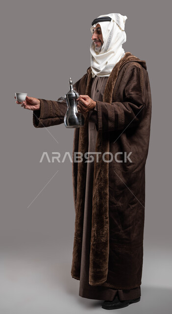 عادات وتقاليد أصيلة في كرم الضيافة، حسن استقبال وإكرام الضيوف، بورتريه لسيد عربي سعودي خليجي مبتسم يرتدي الفروة والغترة التقليدية يقوم بتقديم القهوة العربية الساخنة بإيماءات السرور والترحيب، صورة الجسم بالكامل، خلفية رمادية.
