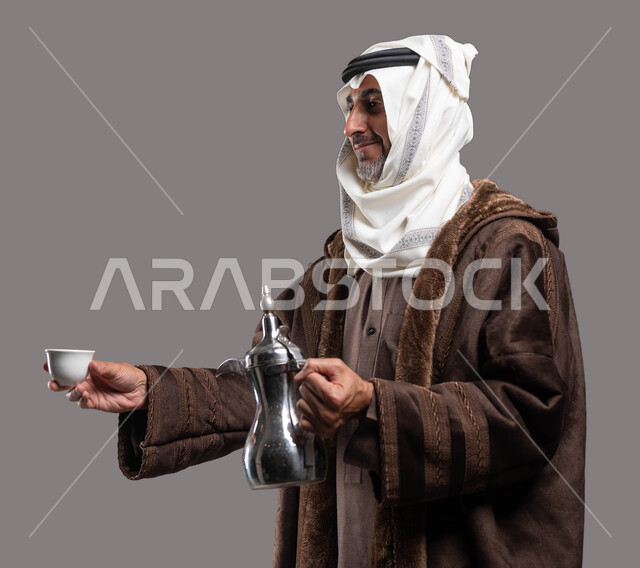استقبال وإكرام الضيوف وفق العادات والتقاليد الأصيلة المتوارثة، أساليب عريقة في كرم الضيافة، الاستمتاع بتناول المشروبات الساخنة في الشتاء، بورتريه مقرب لسيد عربي سعودي خليجي مبتسم يرتدي الفروة والغترة التقليدية يقوم بتقديم القهوة العربية، خلفية رمادية.