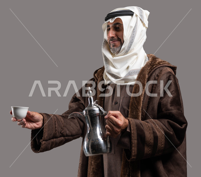 أساليب عريقة في كرم الضيافة، الاستمتاع بتناول المشروبات الساخنة في الشتاء، حسن استقبال وإكرام الضيوف وفق العادات والتقاليد الأصيلة المتوارثة، بورتريه مقرب لسيد عربي سعودي خليجي مبتسم يرتدي الفروة والغترة التقليدية يقوم بتقديم القهوة العربية، خلفية رمادية.