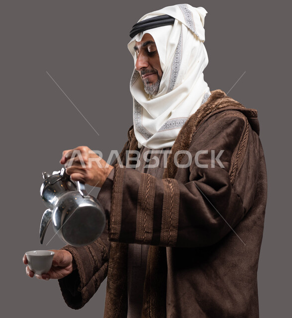 حسن استقبال وإكرام الضيوف وفق العادات والتقاليد الأصيلة المتوارثة، بورتريه مقرب لسيد عربي سعودي خليجي مبتسم يرتدي الفروة والغترة التقليدية يقوم بسكب القهوة العربية ، الاستمتاع بتناول المشروبات الساخنة في الشتاء، خلفية رمادية.