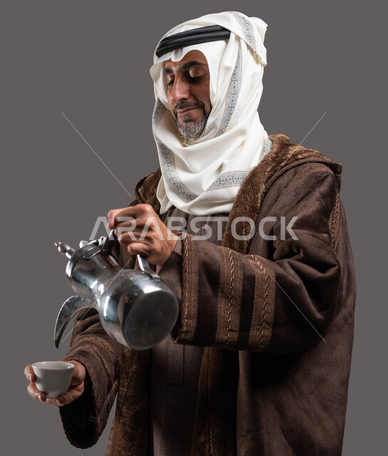 الاستمتاع بتناول المشروبات الساخنة في الشتاء، حسن استقبال وإكرام الضيوف وفق العادات والتقاليد الأصيلة المتوارثة، بورتريه مقرب لسيد عربي سعودي خليجي مبتسم يرتدي الفروة والغترة التقليدية يقوم بسكب القهوة العربية، خلفية رمادية.