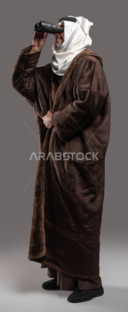 رؤية الأشياء البعيدة، حب الاستكشاف والمغامرة، بورتريه لسيد عربي سعودي خليجي مبتسم يرتدي الفروة والغترة التقليدية يقوم بالنظر من خلال المنظار ثنائي العينين(التلسكوب)، رؤى مستقبلية عند الراشدين والكبار بالسن، صورة الجسم بالكامل، خلفية رمادية.