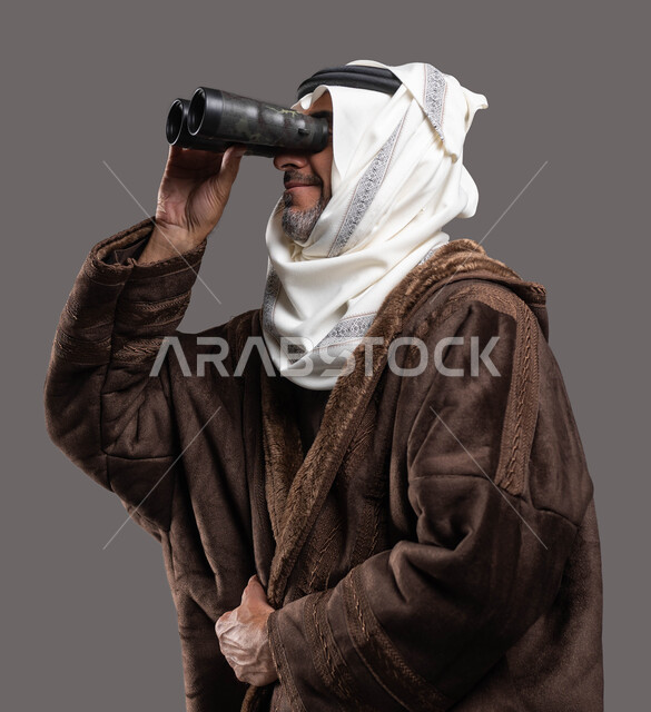 رؤى مستقبلية عند الراشدين والكبار بالسن، رؤية الأشياء البعيدة، حب الاستكشاف والمغامرة، بورتريه مقرب لسيد عربي سعودي خليجي مبتسم يرتدي الفروة والغترة التقليدية يقوم بالنظر من خلال المنظار ثنائي العينين(التلسكوب)، خلفية رمادية.