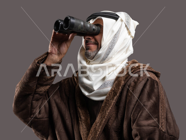 النظر من خلال المنظار ثنائي العينين(التلسكوب)، رؤى مستقبلية عند الراشدين والكبار بالسن، حب الاستكشاف والمغامرة، بورتريه مقرب لسيد عربي سعودي خليجي مبتسم يرتدي الفروة والغترة التقليدية يقوم برؤية الأشياء البعيدة، خلفية رمادية.