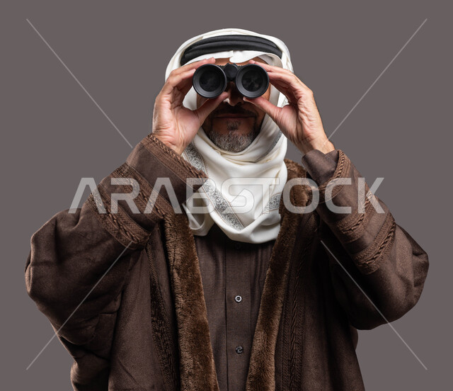 الرحلات الاستكشافية والمغامرة والتخطيط للمستقبل، بورتريه مقرب لرجل شايب عربي سعودي خليجي يرتدي الفروة الشتوية السميكة ويقوم بالنظرمن خلال المنظار الثنائي (التلسكوب)، تخييم في فصل الشتاء، خلفية رمادية.