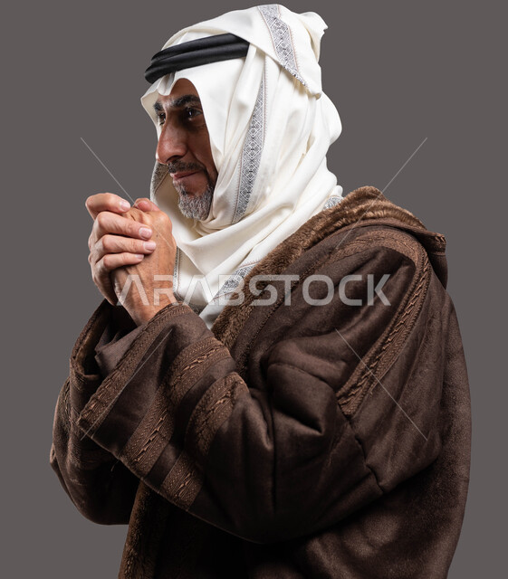 البرد القارس لفصل الشتاء، بورتريه مقرب من الجانب لرجل شايب سعودي خليجي يرتدي الفروة الرجاليه الشتويه والغترة يضم كفيه من شدة البرودة، ايماءات الشعور بالبرد القارس، ملابس مناسبة لفصل الشتاء،أزياء سعودية تقليدية، خلفية رمادية .