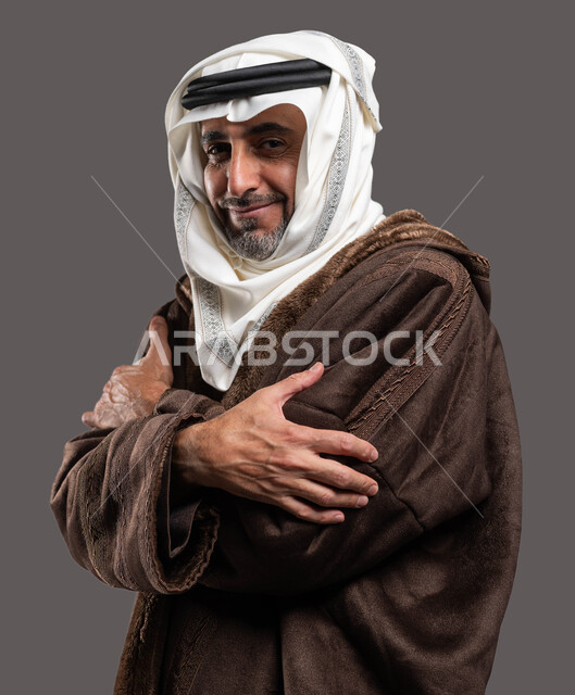 الإحساس الشديد بالبرد في الأجواء الشتوية، بورتريه من الجانب مقرب لرجل مسن عربي خليجي سعودي يرتدي الفروة الرجالية السميكة ويضع الغترة التقليدية يقف بأيدي متقاطعة، إيماءات الشعور بالصقيع والقشعريرة، خلفية رمادية.