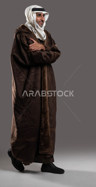 كثرة الحركة لتقليل من برودة الجسم في فصل الشتاء، بوتريه لرجل مسن عربي خليجي سعودي خليجي يرتدي الفروه الشتوية السميكة ويضع العقال والغترة السعودية يمشي بايدي مكتفة، ايماءات الشعور الشديد بالبرد، صورة لكامل الجسم، خلفية رمادية.