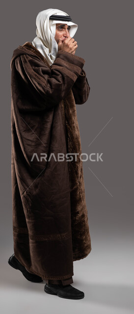 المشي و الحركة للحفاظ على دفئ الجسم في الاجواء الشتوية، بورتريه لرجل شايب عربي سعودي خليجي يرتدي الفروة الشتوية الدافئة ويضع العقال والغترة يقوم بالتنفس العميق في يديه لتدفئتها، إيماءات الشعور بالبرد الشديد، صورة الجسم بالكامل، خلفية رمادية.