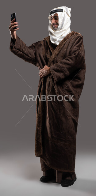 هواية التصوير باستخدام الجوال المحمول لصنع ذكريات جميلة، بورتريه لرجل عربي سعودي خليجي برتدي اللباس الشتوي الدافئ ويضع العقال والغترة التقليدية يقوم بالتقاط صورة سيلفي عبر الهاتف المحمول، صورة لكامل الجسم، إجراء بث مباشر، إيماءات الشعور بالبرد، خلفية رمادية.