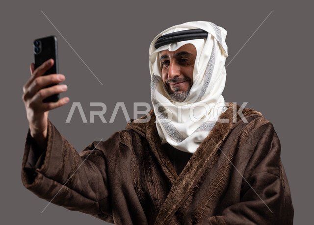توثيق اللحظات المميزة باستخدام الهاتف المحمول، بورتريه مقرب لرجل عربي خليجي سعودي مبتسم يرتدي الفروة  و يضع الغترة والعقال يقوم بالتقاط صورة سيلفي، التواصل مع الاهل و الاصدقاء بمكالمة فيديو، ايماءات الاستمتاع بالاجواء الشتوية، خلفية رمادية.