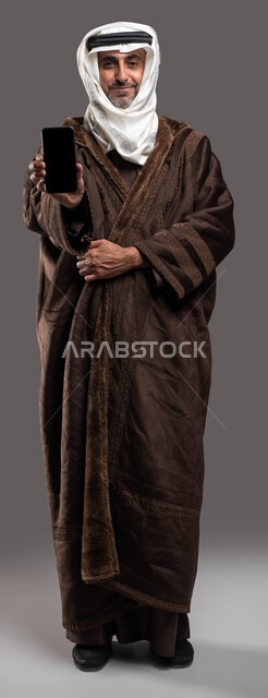 The contribution of mobile phone applications and services to facilitating the purchase of winter clothes, a portrait of an elderly Saudi Arabian Gulf man wearing a winter fur, wearing an iqal and a ghutra, holding a mobile phone in his hand with a blank black screen, full-length body photo, gray background.