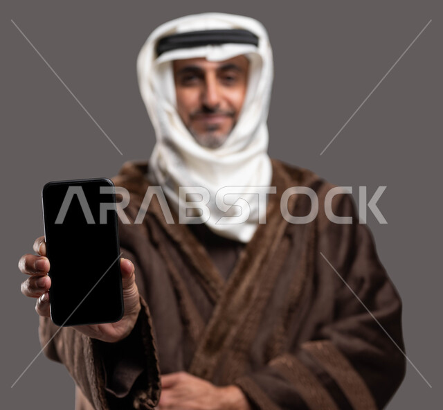 توفر تطبيقات الهاتف المحمول، التسوق السريع للملابس الشتوية، بورتريه مقرب لرجل مسن عربي سعودي خليجي يرتدي االفروة الشتوية ويضع العقال والغترة يحمل بيده الجوال بشاشة سوداء فارغة، إيماءات الشعور بالبرد، أهمية التكنولوجيا في التسويق و الاعلان، خلفية رمادية .