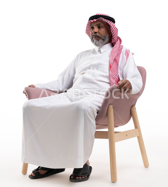 إيماءات التفكير وشرود الزهن، الشعور بالتعب والإرهاق، بورتريه لرجل كبير بالسن عربي سعودي خليجي يرتدي الشماغ والثوب التقليدي يجلس على كرسي مريح وينظر إلى شيء ما، خلفية بيضاء
