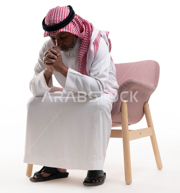 الشعور بالتعب والإرهاق، التفكير والقلق بإيماءات الحزن لخسارة شيء ما، حل المشكلات واتخاذ القرارات، بورتريه لرجل كبير بالسن عربي سعودي خليجي يرتدي الشماغ والثوب التقليدي يجلس على كرسي مريح يضع يديه على وجهه، خلفية بيضاء