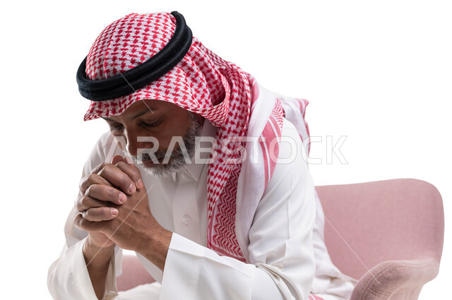التفكير والقلق بإيماءات الحزن لخسارة شيء ما، الشعور بالتعب والإرهاق، حل المشكلات واتخاذ القرارات، بورتريه مقرب لرجل كبير بالسن عربي سعودي خليجي يرتدي الشماغ والثوب التقليدي يجلس على كرسي مريح يضع يديه على وجهه، خلفية بيضاء