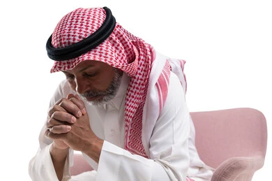 Thinking and worrying with gestures of sadness for losing something, feeling tired and exhausted, solving problems and making decisions, close-up portrait of an elderly Saudi Arabian Gulf man wearing a shemagh and traditional thobe, sitting in a comfortable chair with his hands on his face, white background.