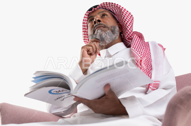 محو الأمية والتعلم والتثقيف، قضاء أوقات الفراغ بالقراءة والمطالعة، بورتريه مقرب لرجل كبير بالسن عربي سعودي خليجي يرتدي الشماغ والثوب التقليدي يحمل بيده كتاب علمي تثقيفي ينظر إلى الأعلى بإيماءات التفكير وشرود الذهن، خلفية بيضاء
