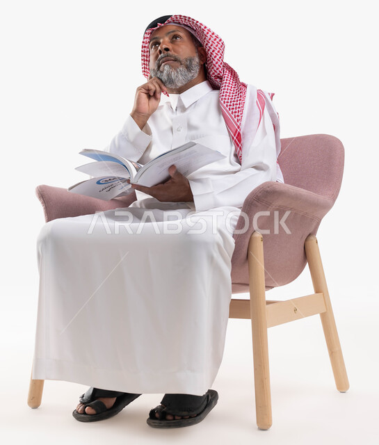 مفهوم محو الأمية والتعلم، قضاء أوقات الفراغ بالقراءة والمطالعة، بورتريه لرجل كبير بالسن عربي سعودي خليجي يرتدي الشماغ والثوب التقليدي يحمل بيده كتاب علمي تثقيفي ينظر إلى الأعلى بإيماءات التفكير وشرود الذهن، خلفية بيضاء