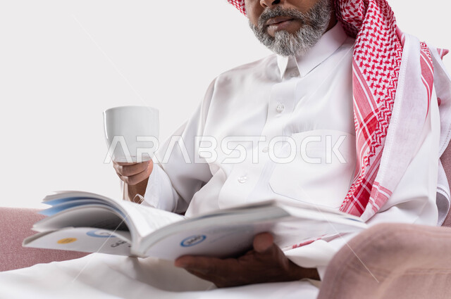 الاستمتاع بتناول المشروبات الساخنة، التزود بالعلم والمعرفة، قضاء وقت الفراغ بالتثقيف والتعلم، مفهوم محو الأمية، بورتريه مقرب  لرجل كبير بالسن عربي سعودي خليجي يرتدي الشماغ والثوب التقليدي يشرب القهوة ويمارس هواية قراءة الكتب المفيدة في وقت الفراغ، خلفية بيضاء
