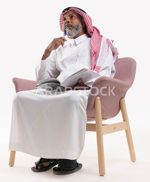 قضاء وقت الفراغ بالقراءة والمطالعة، مفهوم محو الأمية والتعلم، بورتريه لرجل كبير بالسن عربي سعودي خليجي يرتدي الشماغ والثوب التقليدي يحمل بيده كتاب علمي تثقيفي ينظر إلى الأعلى بإيماءات التفكير وشرود الذهن، خلفية بيضاء