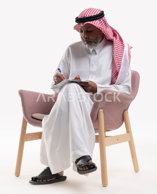 ايماءات تدل على الاندماج والتمعن، حل المشكلات واتخاذ القرارات، تدوين وتسجيل الملاحظات، بورتريه مقرب لرجل عربي سعودي خليجي كبير بالسن يرتدي الشماغ والثوب التقليدي يقوم بكتابة المعلومات على المفكرة الورقية، خلفيه بيضاء