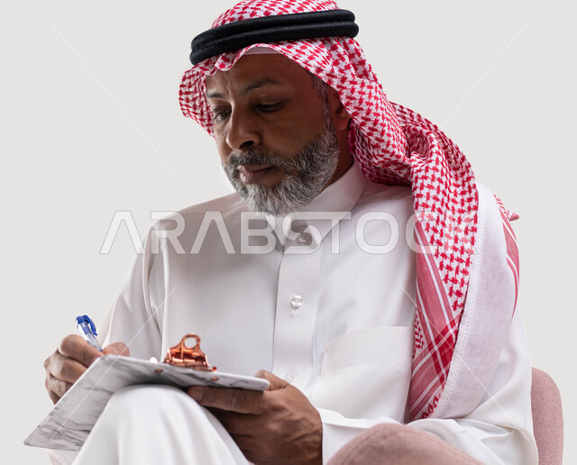 حل المشكلات واتخاذ القرارات، تدوين وتسجيل الملاحظات، تعابير وايماءات تدل على الاندماج والتمعن، بورتريه مقرب لرجل عربي سعودي خليجي كبير بالسن يرتدي الشماغ والثوب التقليدي يقوم بكتابة المعلومات على المفكرة الورقية، خلفيه بيضاء