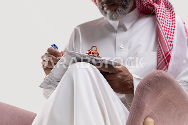 تدوين وتسجيل الملاحظات، تعابير وايماءات تدل على الاندماج والتمعن، بورتريه مقرب لرجل عربي سعودي خليجي كبير بالسن يرتدي الشماغ والثوب التقليدي يجلس على كرسي مريح يقوم بكتابة المعلومات على المفكرة الورقية باستخدام القلم، خلفيه بيضاء