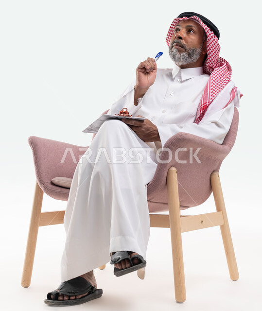كتابة وتسجيل الملاحظات، تعابير التمعن والتركيز، بورتريه لرجل عربي سعودي خليجي كبير بالسن يرتدي الشماغ والثوب التقليدي يجلس على كرسي مريح يمسك بيديه مفكرة وقلم، النظر الى شيء ما بإيماءات التفكير، خلفيه بيضاء