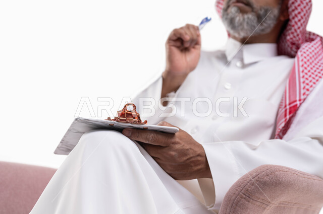 تعابير وايماءات التمعن والتركيز، كتابة وتسجيل الملاحظات، بورتريه مقرب لرجل عربي سعودي خليجي كبير بالسن يرتدي الشماغ والثوب التقليدي يجلس على كرسي مريح يمسك بيديه مفكرة وقلم، خلفيه بيضاء