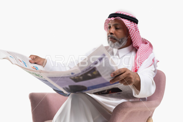 الإطلاع على المقالات والأحداث العالمية من خلال الصحائف, تصفح الجرائد ومتابعة أحدث الأخبار، ممارسة هواية القراءة, بورتريه مقرب لرجل عربي خليجي سعودي كبير بالسن يرتدي الشماغ والثوب التقليدي يجلس على كرسي مريح يقوم بقراءة جريدة ورقيه، خلفية بيضاء