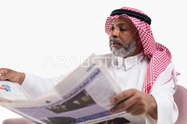 تصفح الجرائد ومتابعة أحدث الأخبار، الإطلاع على المقالات والأحداث العالمية من خلال الصحائف, ممارسة هواية القراءة, بورتريه مقرب لرجل عربي خليجي سعودي كبير بالسن يرتدي الشماغ والثوب التقليدي يجلس على كرسي مريح يقوم بقراءة جريدة ورقيه، خلفية بيضاء