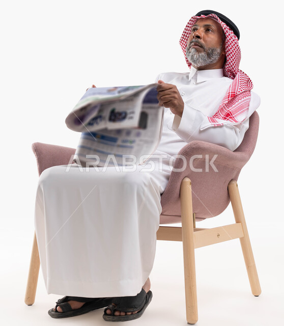متابعة أحدث الأخبار من خلال تصفح الجرائد، ممارسة هواية القراءة, الإطلاع على المقالات والأحداث العالمية من خلال الصحائف, بورتريه لرجل عربي خليجي سعودي كبير بالسن يرتدي الشماغ والثوب التقليدي يجلس على كرسي مريح يمسك جريدة ورقية ينظر الى شيء ما، خلفية بيضاء
