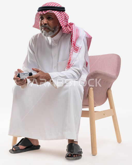 الاندماج والتفاعل مع ألعاب البلايستيشن، بورتريه لرجل عربي سعودي خليجي كبير بالسن يرتدي الشماغ والثوب التقليدي يجلس على كرسي مريح يحمل بيده عصا التحكم يقوم باللعب في ألعاب الفيديو، اجهزة وتقنيات الكترونية حديثة ترفيهيه, قضاء وقت ممتع ومسلي باللعب، خلفية بيضاء